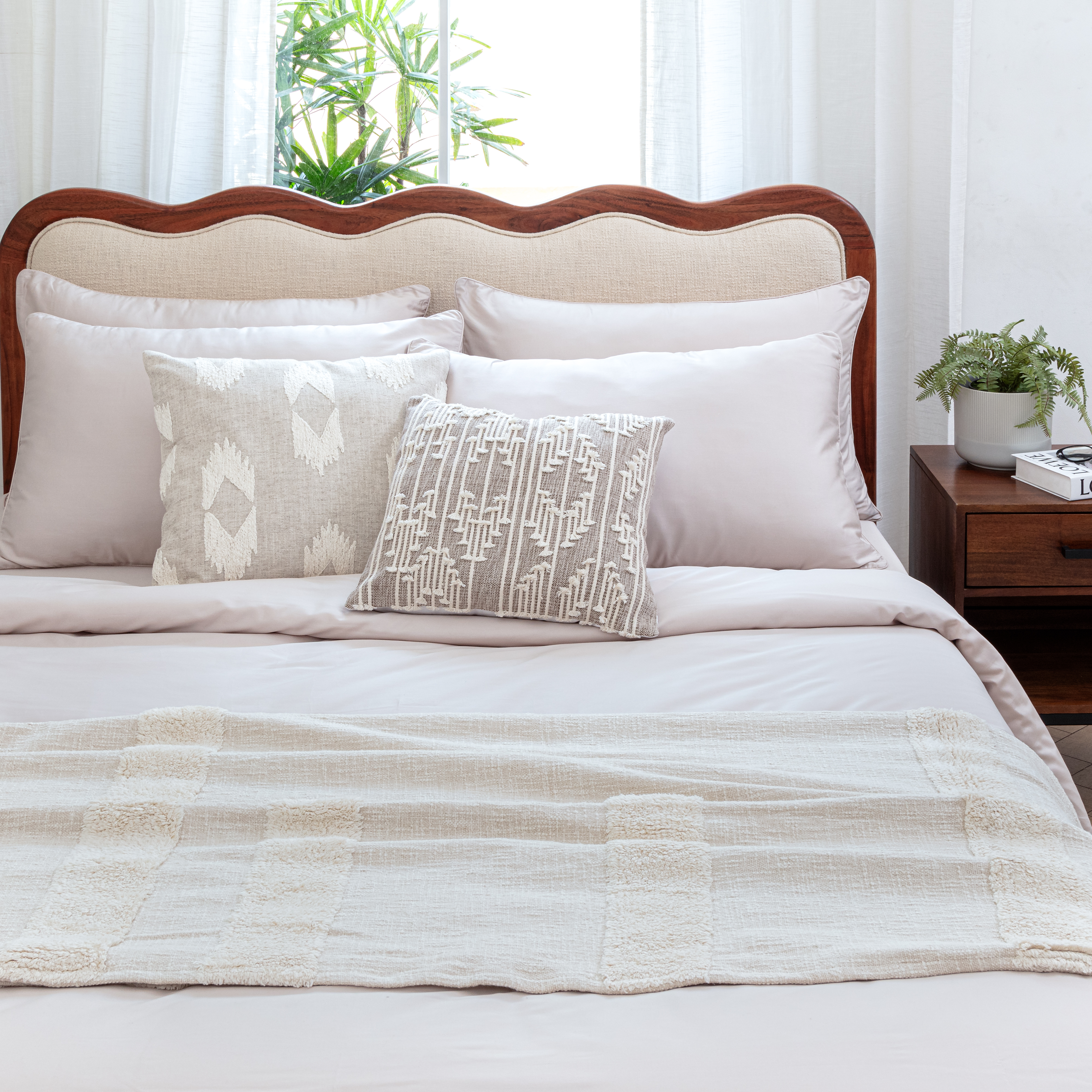 A bed with sand coloured high thread count cotton bed sheets layered with a throw and decorative cushions