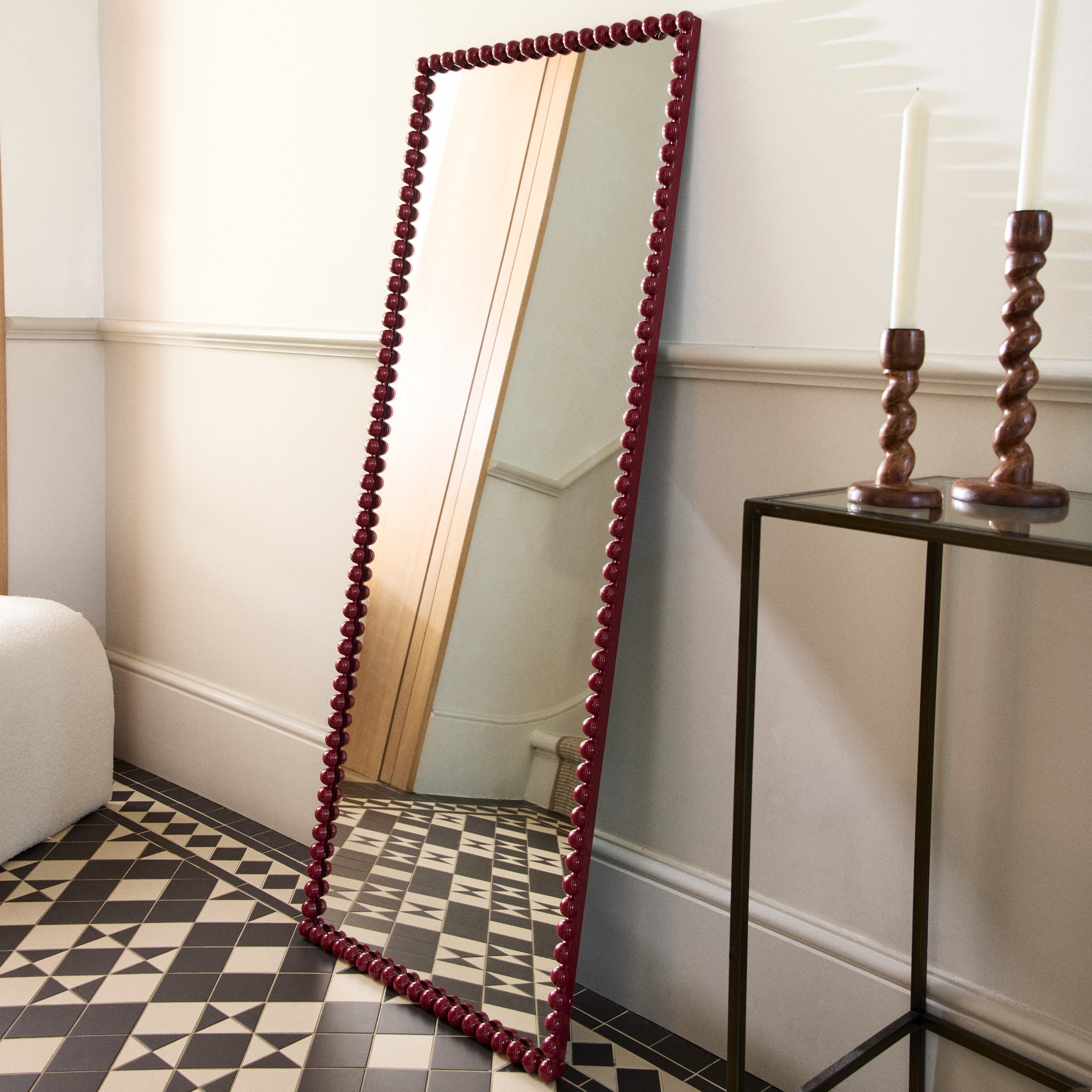 Tall red bobbin mirror against a wall in a hallway next to wood sculpted candle holders